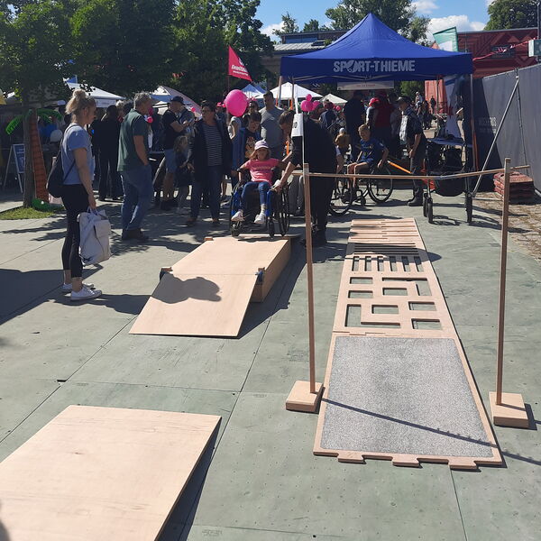 Rollstuhlparcours, bestehend aus Rampen, Wippe und unebenen Platten, gefertigt aus Holz. Kinder die Hürden im Rollstuhl überwinden. Festival-Treiben.