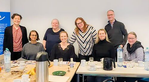 In einem Raum. Tisch gedeckt mit Kaffee, Wasser. 4 Personen sitzen am Tisch, 4 Personen stehen versetzt dahinter. 7 Frauen, ein Mann, alle lächeln in die Kamera.