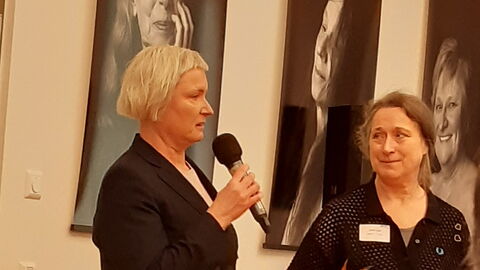 2 Frauen stehen vor großen schwarz-weis- Portraits von Frauen. Sie tragen schwarze Kleidung und eröffnen die Veranstaltung.