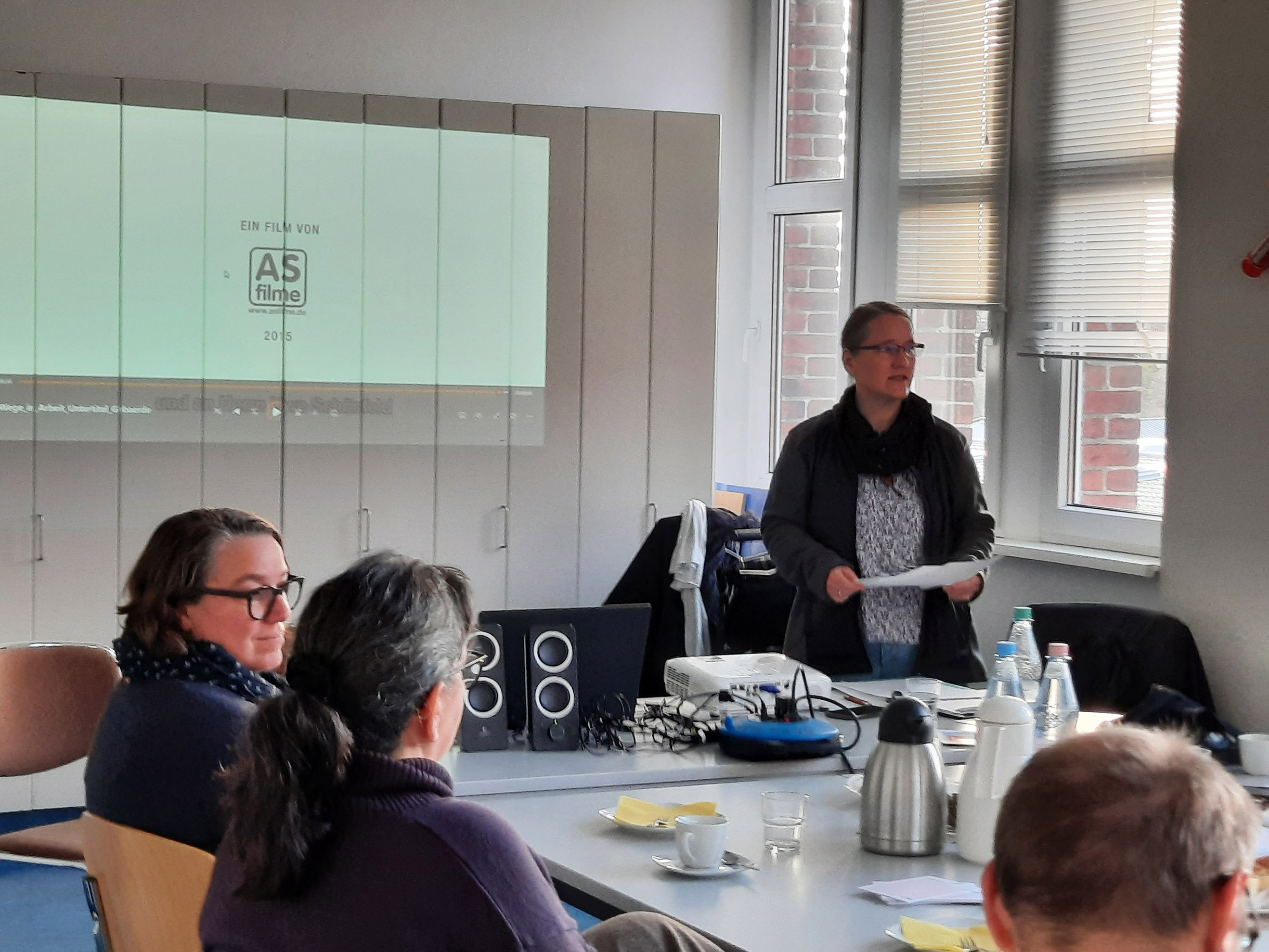 Konferenzraum. Zwei Frauen sitzen am Tisch. Eine Frau steht vorne mit Notizen in den Händen und spricht. Auf dem Tisch sind Getränke und Geschirr.