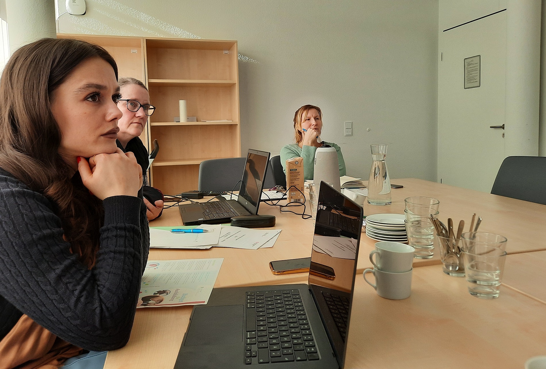 Großer Raum, großer Tisch, 2 Frauen mit Laptops vor sich und eine Frau mit Stift und Notizheft. Auf dem Tisch, Tassen, Teller, Wasserkaraffe und Zettel und Stifte.
