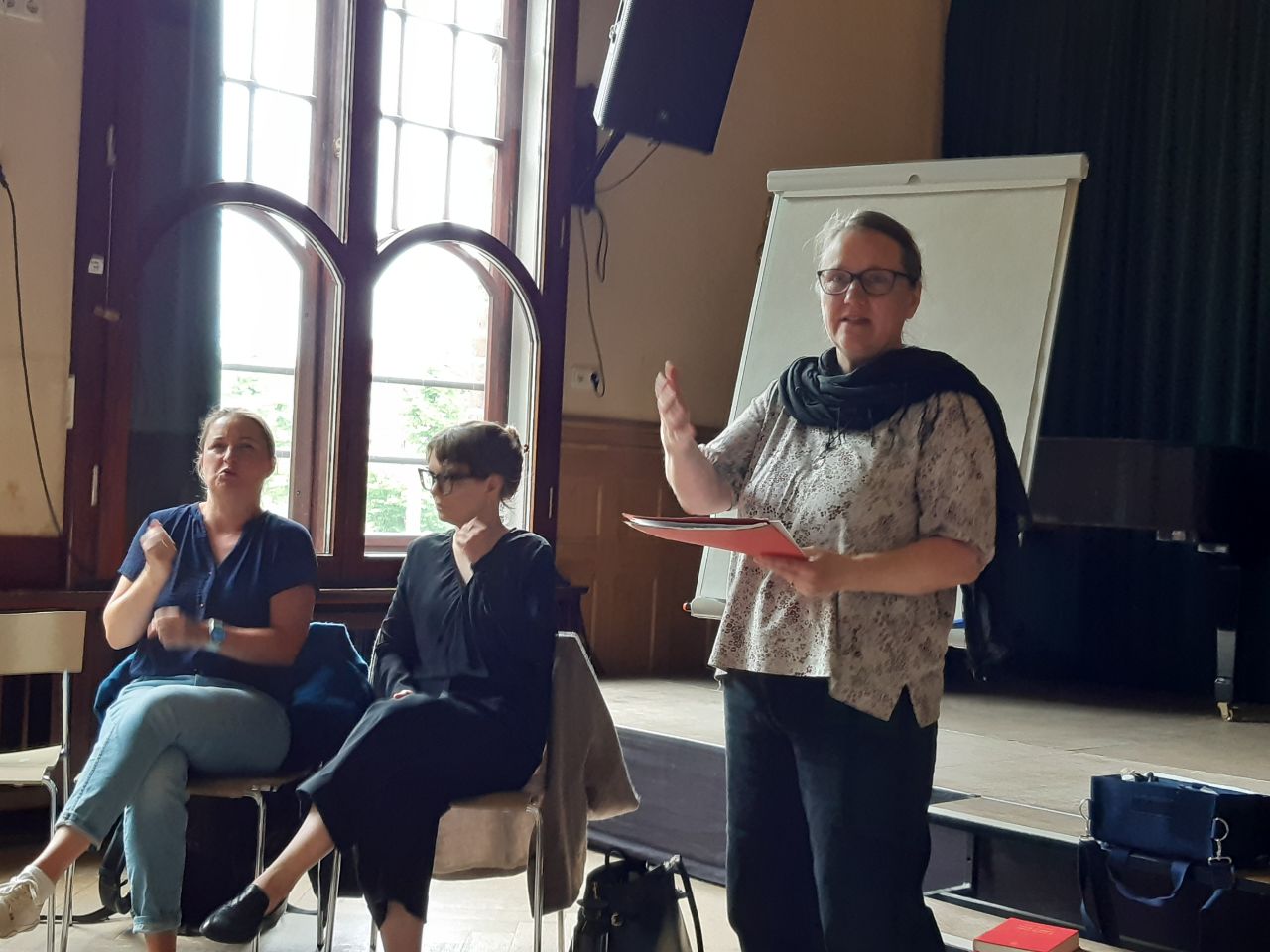 Frau steht vor einem Flipchart und spricht, in der Hand hält sie einen Hefter. Links neben ihr sitzen zwei Frauen in dunkler Kleidung und übersetzen in Gebärdensprache.
