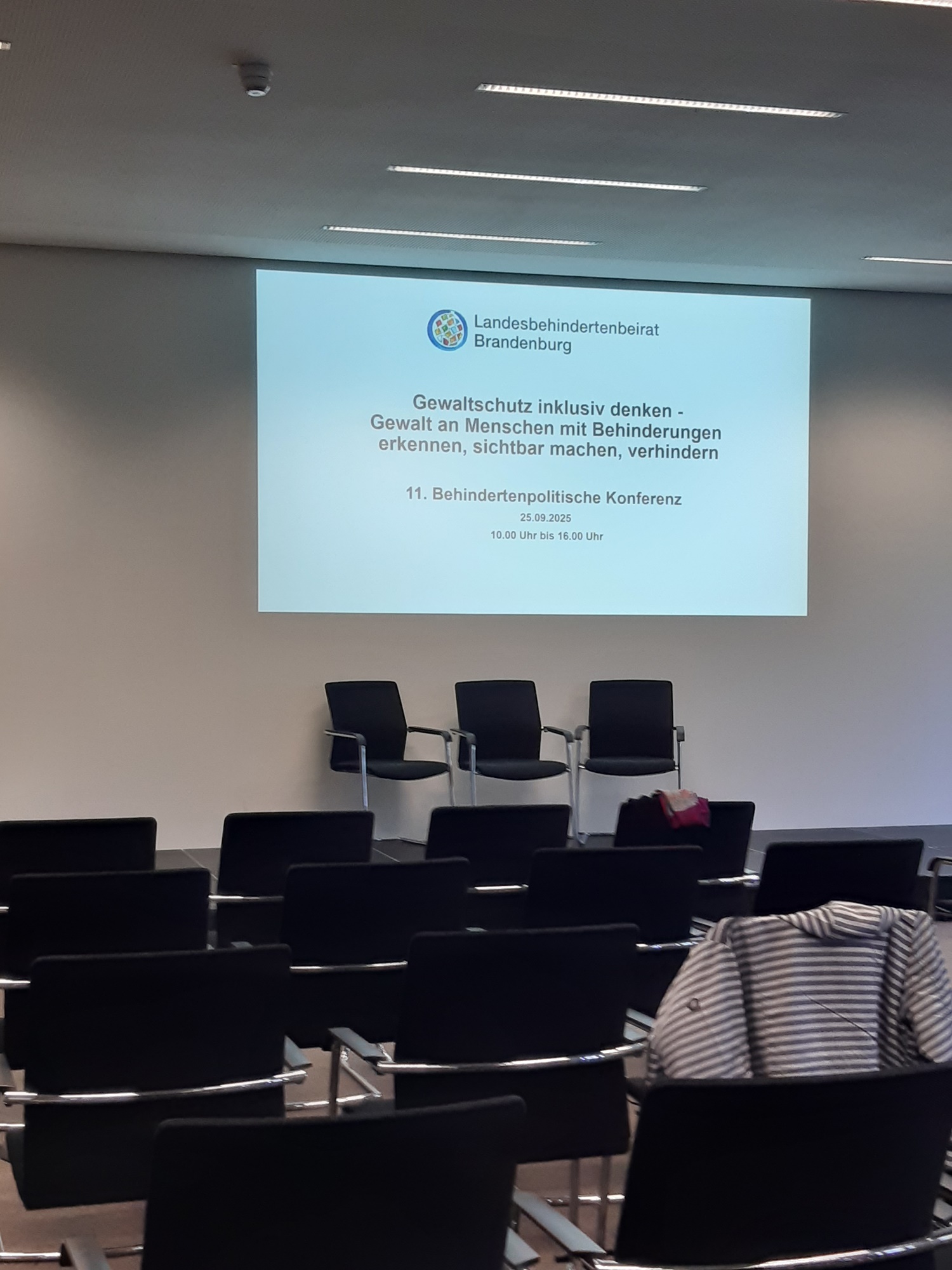 Leerer Konferenzraum, drei Stühle auf einem Podium. Darüber mit Beamer an die Wand projiziert. "Gewaltschutz inklusiv denken - Gewalt an Menschen mit Behinderung erkennen, sichtbar machen, verhindern.