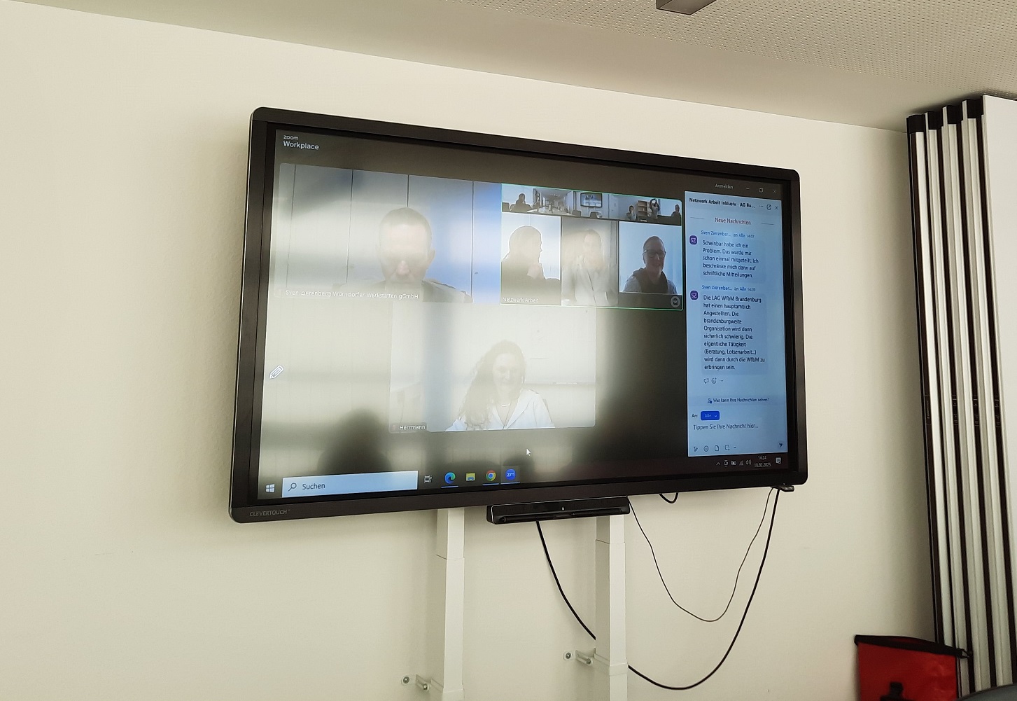 Monitor an der Wand. Ein Online Meeting. Mehrere Menschen sind zugeschaltet.