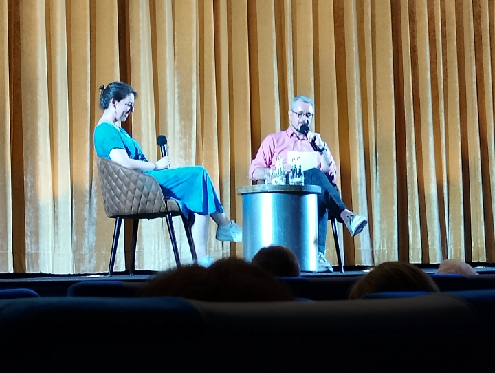 Podiumsgespräch: Eine frau und ein Mann sitzen vor einem gelben Kinovorhang. Beide haben ein Mikro in der Hand. Der Mann spricht.