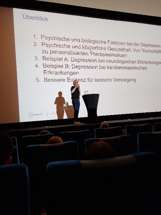Eine Präsentation über Psychische und biologische Faktoren bei Depression, Ein Mann in dunkler Kleidung spricht in ein Mikrofon.