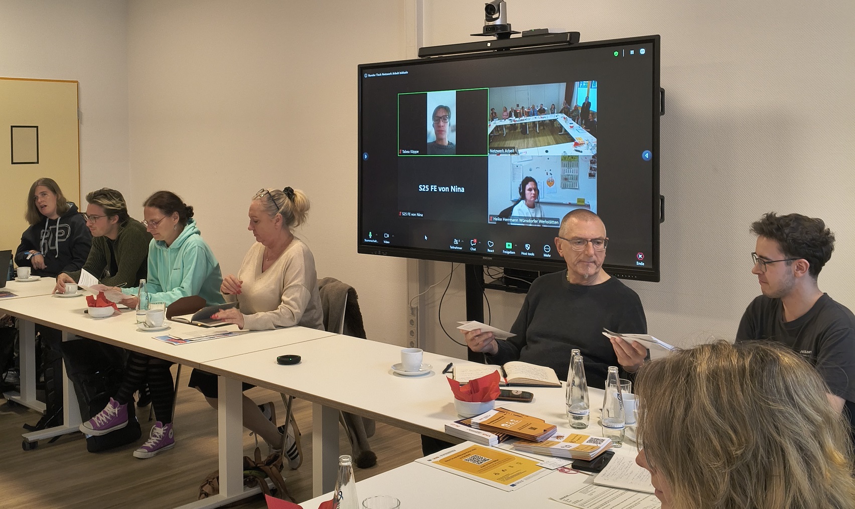 Im Fokus eine Videokonferenz. Großer Monitor an der Wand, darauf Kacheln mit Menschen. Menschen sitzen mit Rücken zum Monitor davor.